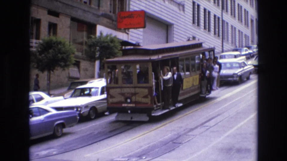1973 San Francisco cable car traveling on a steep street with passengers standing on the side platform, surrounded by 1970s cars and downtown buildings.