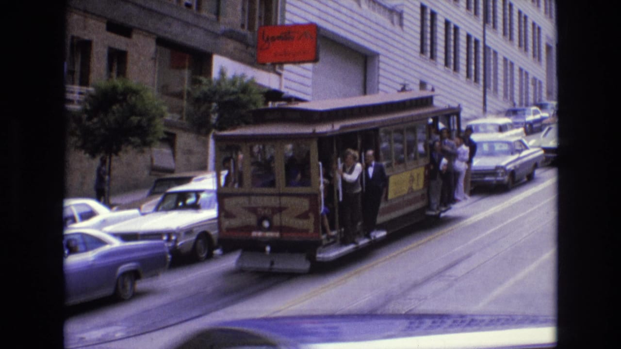 San Francisco cable car in 1973 moving along a steep urban street, with passengers riding on the open side and several period cars lining the roadway.