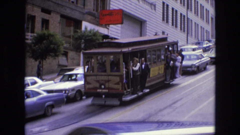 San Francisco cable car in 1973 moving along a steep urban street, with passengers riding on the open side and several period cars lining the roadway.