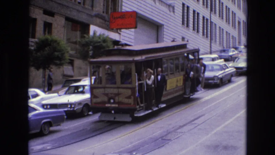 Rear-side view of a 1973 San Francisco cable car on a steep street, with visible passengers, parked sedans, rail tracks, and tall commercial buildings.
