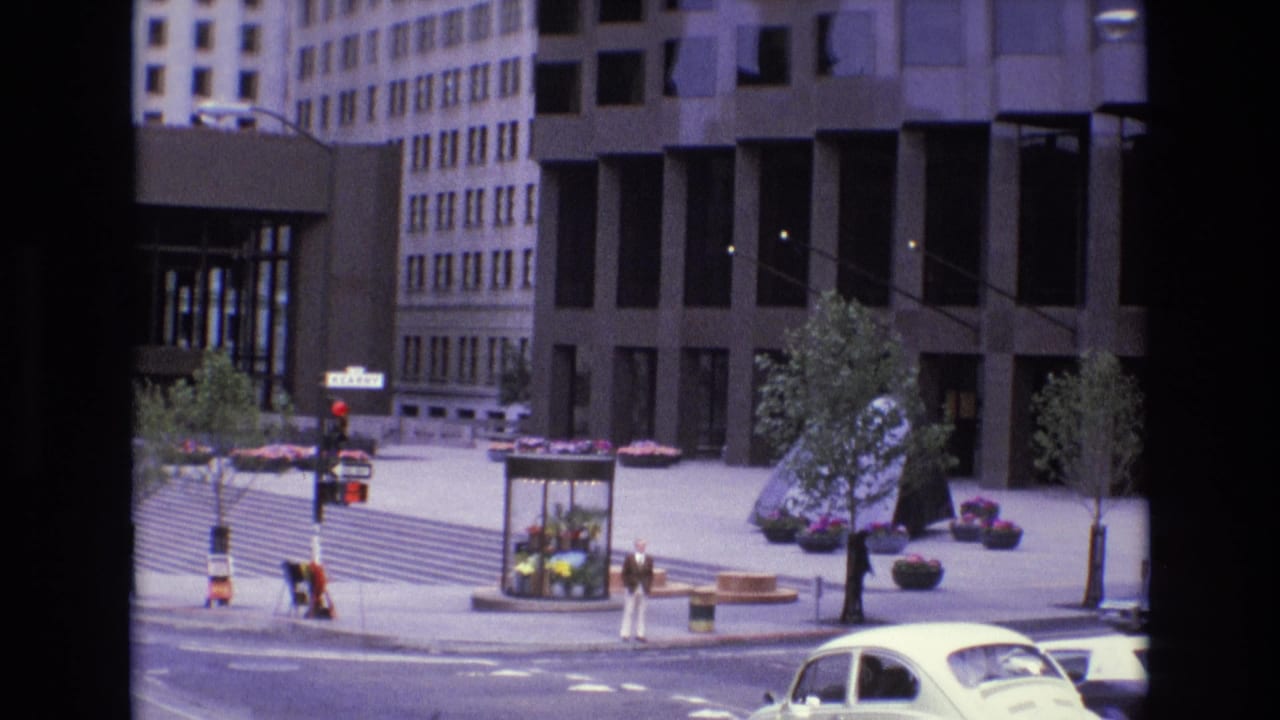 1973 San Francisco street view across a quiet plaza with mid-century office towers, a flower kiosk, small trees, pedestrians, and a white Volkswagen Beetle in the foreground.