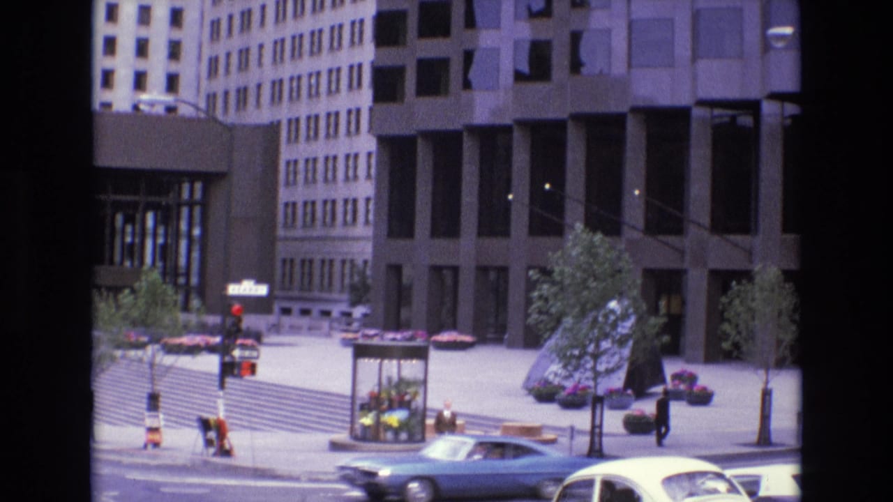 Blue sedan passes in front of a plaza with pedestrians, planters, trees, and dark glass office buildings.
