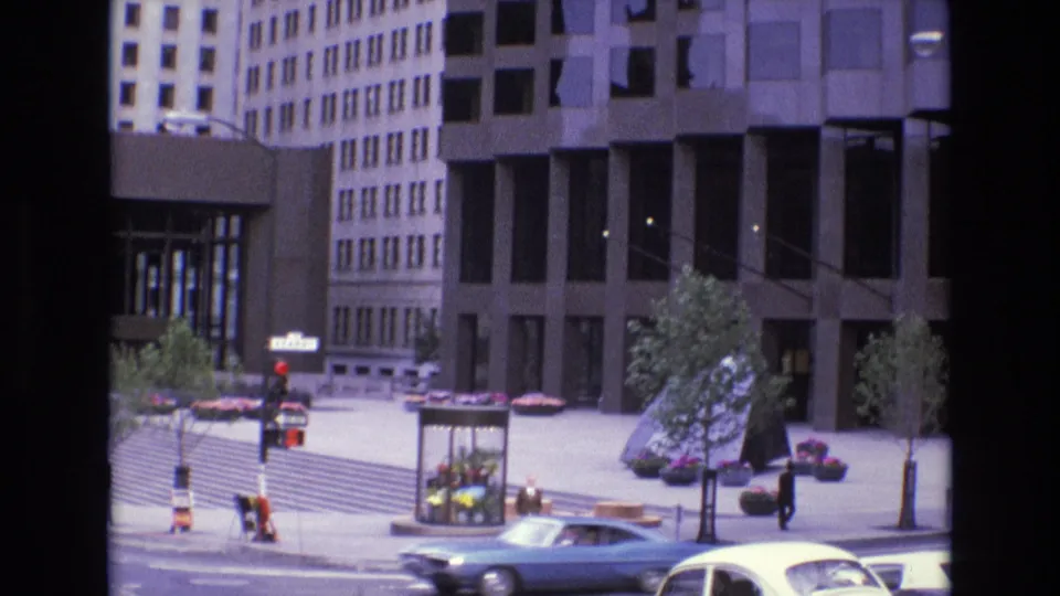 1973 San Francisco plaza scene with a blue sedan crossing the foreground, pedestrians on the pavement, planted trees, flower displays, and mid-century office building facades behind them.