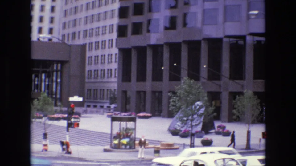 1973 San Francisco 8mm frame showing a broad downtown plaza with pedestrians, a flower kiosk, potted plantings, office towers, and a white car moving along the street edge.