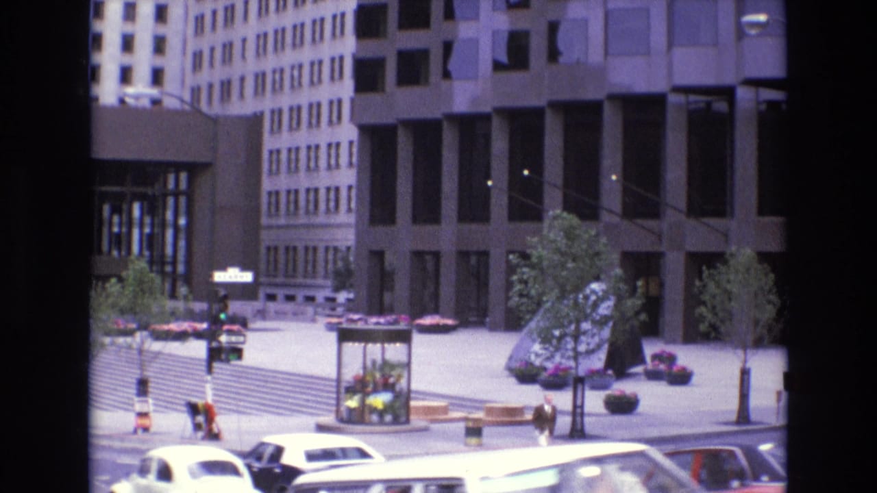 1973 San Francisco street view with mid-century cars passing in front of plaza steps, pedestrians, trees, and modern office buildings.