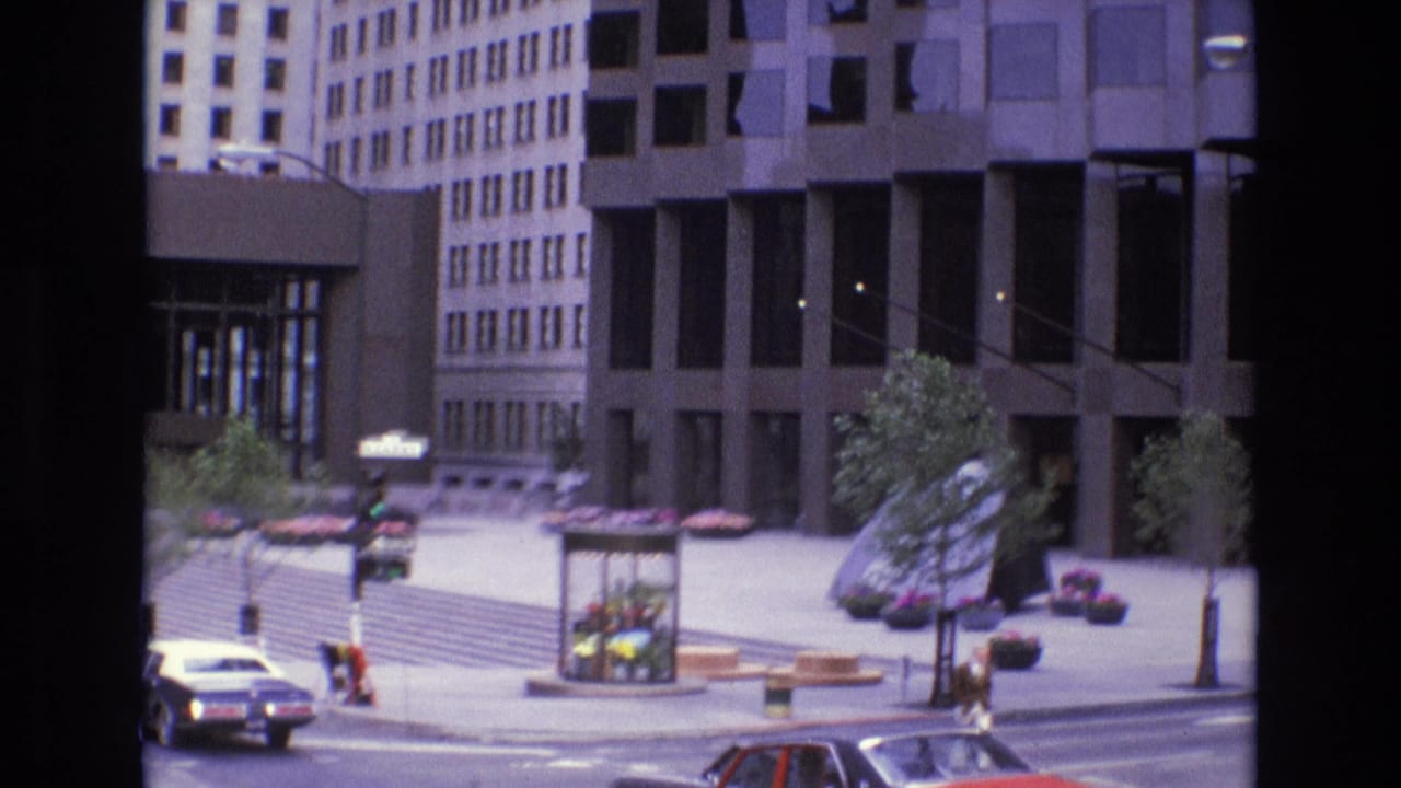 Street corner with cars, pedestrians, modern buildings, trees, and plaza seating.