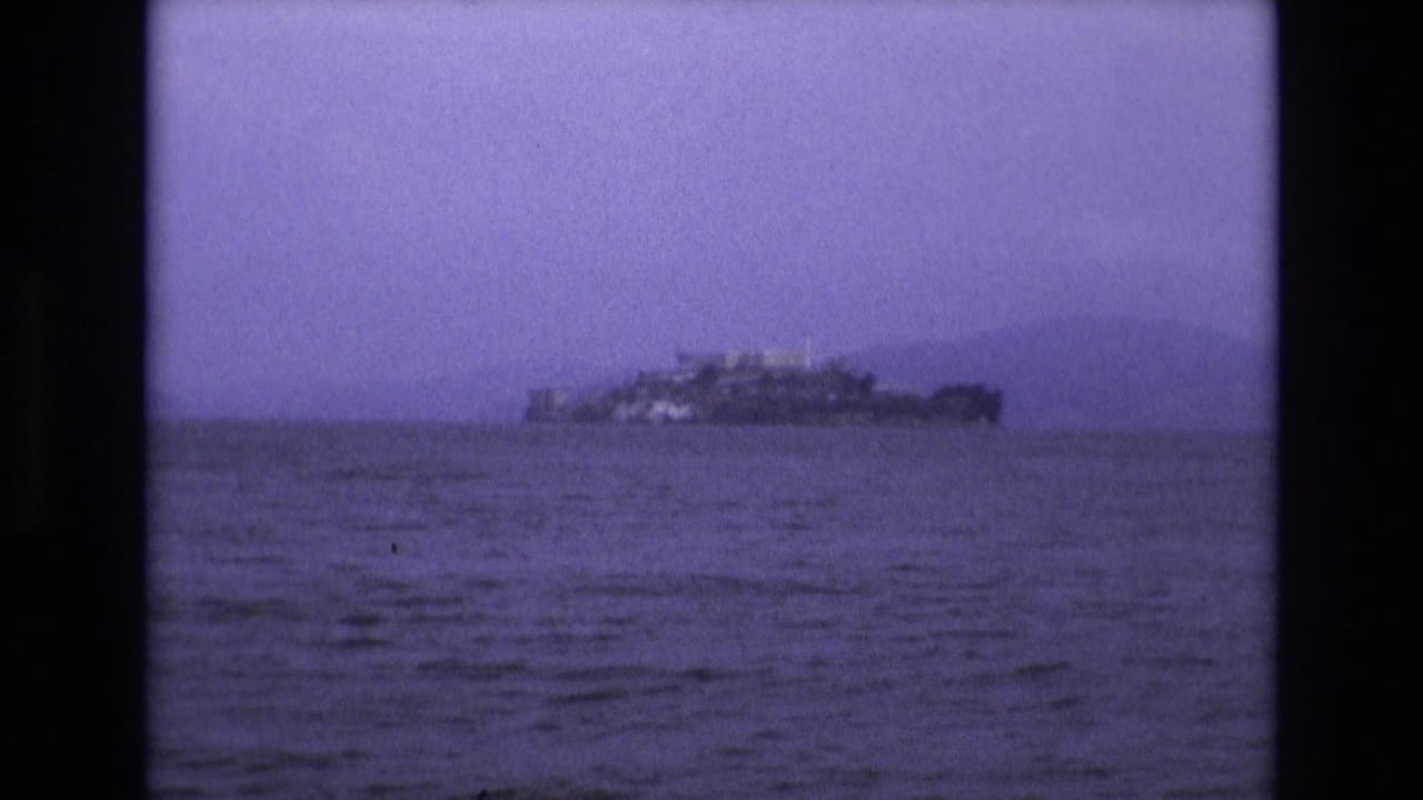 1973 8mm home-movie frame showing Alcatraz Island in the distance across San Francisco Bay, tinted purple with dark film borders.