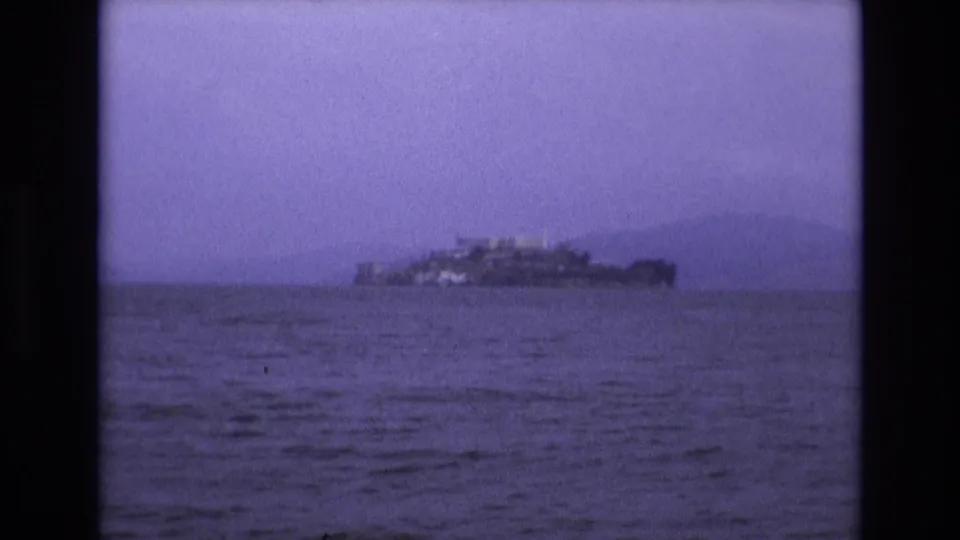 1973 8mm home-movie frame showing Alcatraz Island in the distance across San Francisco Bay, tinted purple with dark film borders.