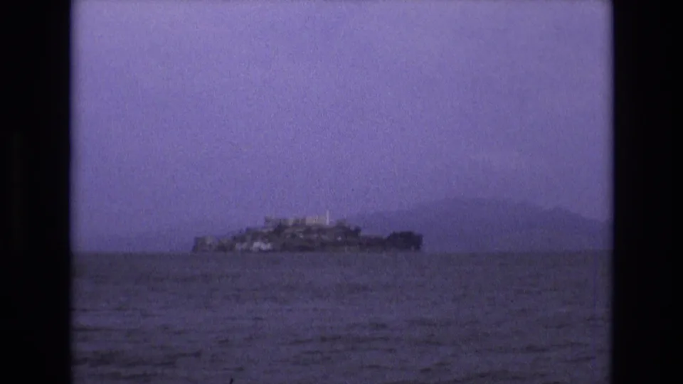 1973 San Francisco Bay home-movie frame with Alcatraz Island low on the horizon, softened by haze and purple film tint.