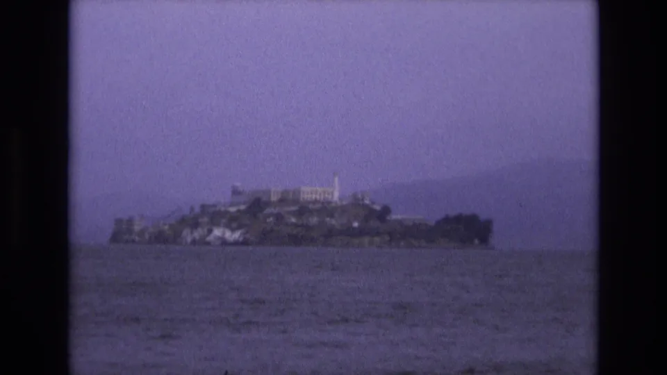 Distant 1973 8mm view of Alcatraz Island in San Francisco Bay, with low buildings and rocky shoreline seen across rippled water.