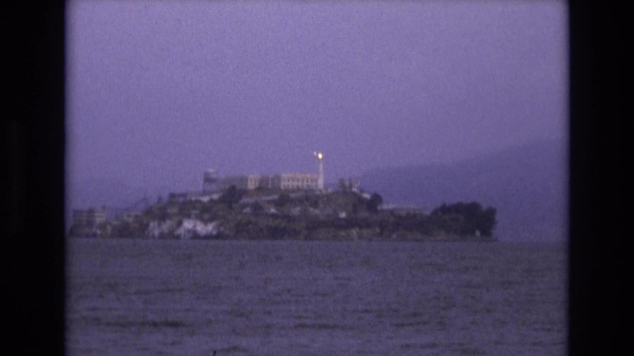 Distant 1973 San Francisco Bay view of Alcatraz Island, with the prison complex and a small bright light visible against a purple-tinted sky.