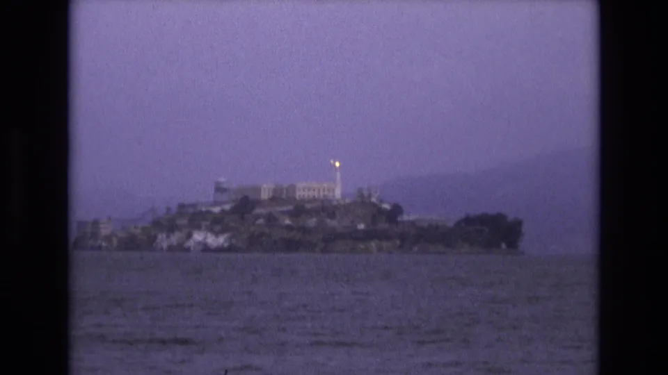 Distant 1973 San Francisco Bay view of Alcatraz Island, with the prison complex and a small bright light visible against a purple-tinted sky.
