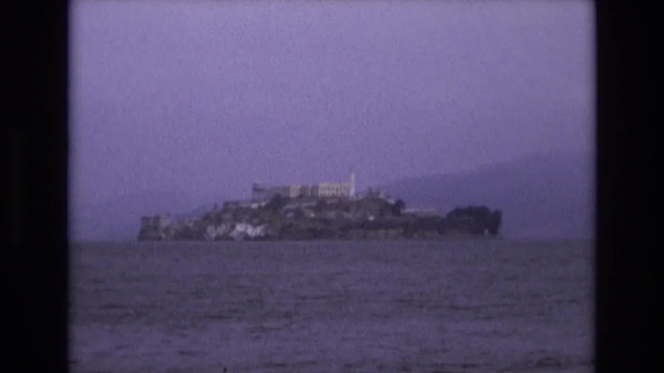 1973 8mm view of Alcatraz Island in San Francisco Bay, seen from a distance with water in the foreground and hazy hills behind.