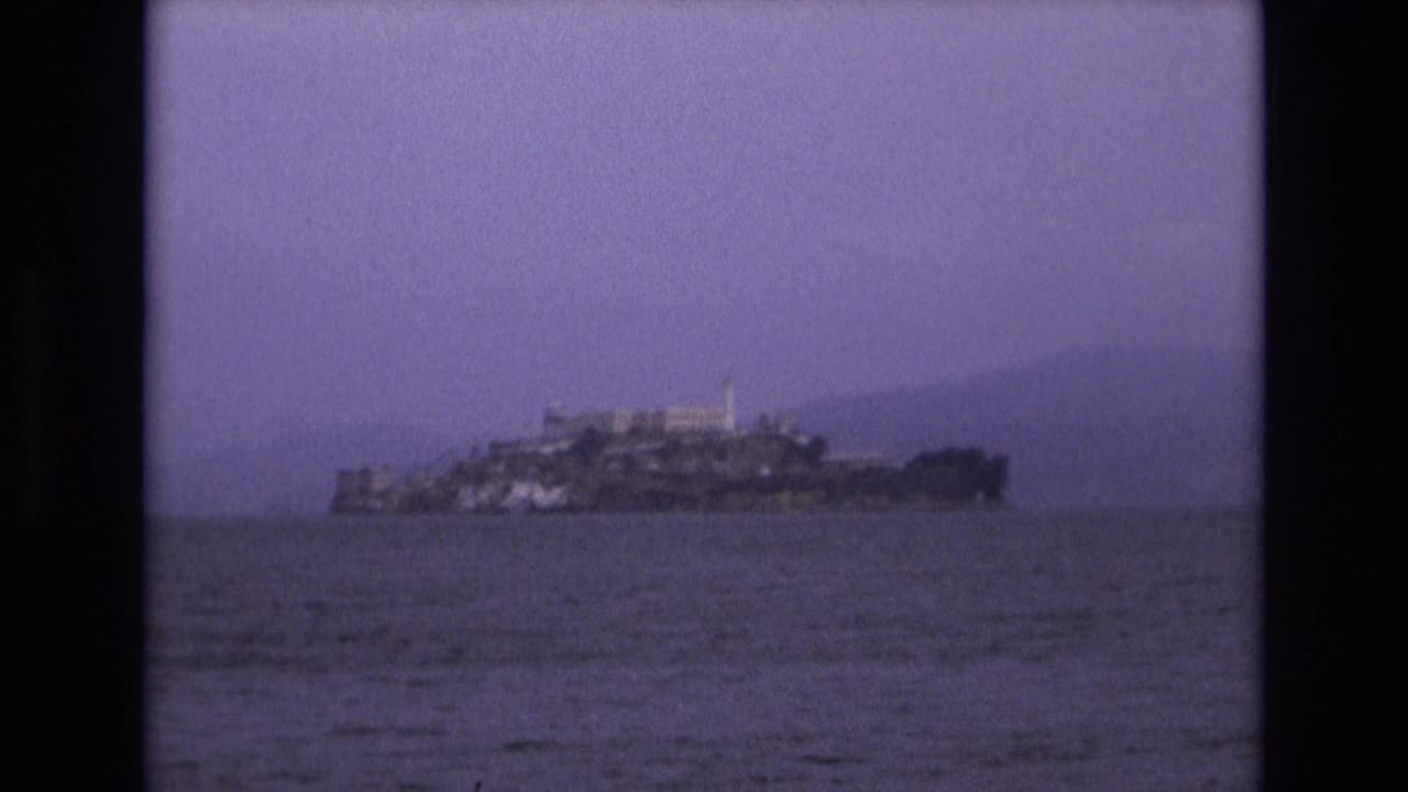 Distant 1973 home-movie frame of Alcatraz Island centered across San Francisco Bay, with soft focus, grain, and pale haze in the background.