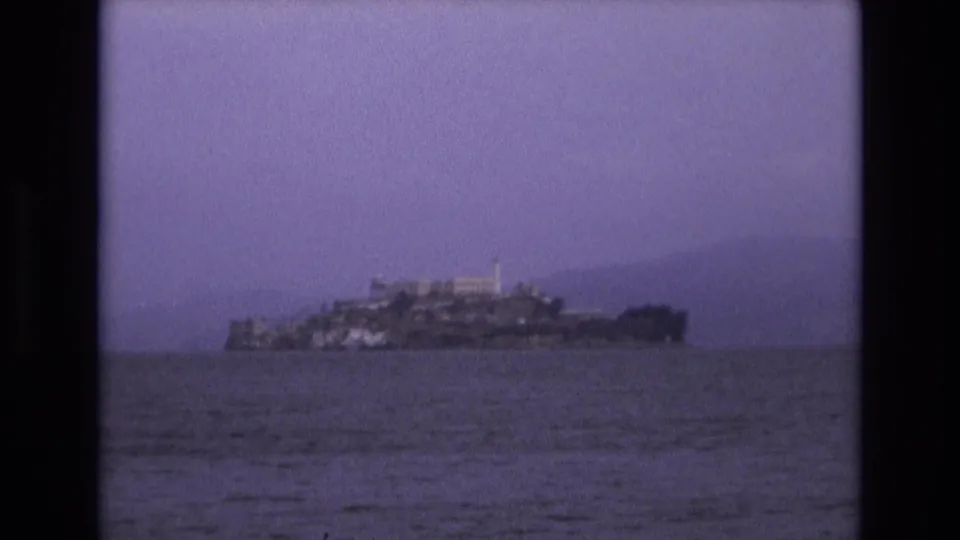 Distant 1973 home-movie frame of Alcatraz Island centered across San Francisco Bay, with soft focus, grain, and pale haze in the background.