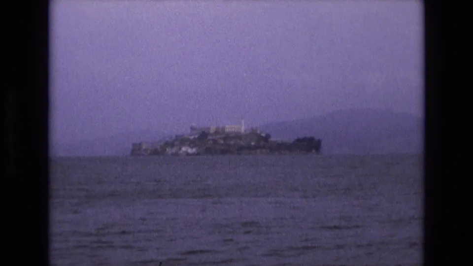 1973 8mm frame showing Alcatraz Island low across the bay, with water occupying the foreground and a purple-tinted, soft-focus film look.