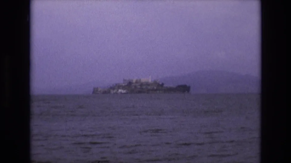 Distant view of Alcatraz Island across San Francisco Bay in 1973 8mm home-movie footage, with soft grain, purple tint, and open water in the foreground.