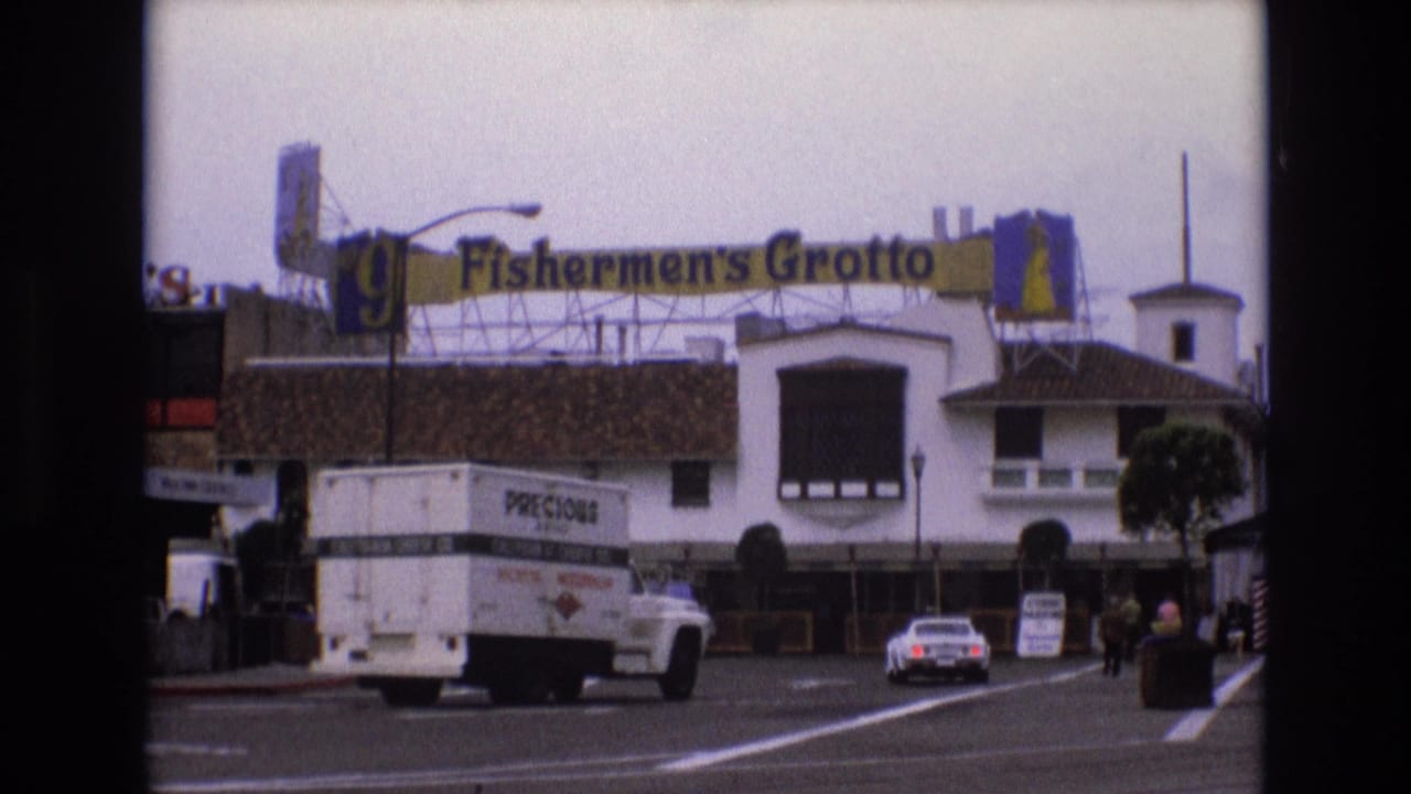 White delivery truck and white car pass Fishermen's Grotto storefronts on a city street.