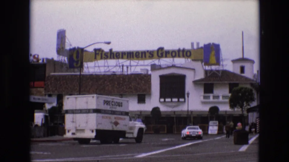 1973 San Francisco home-movie frame showing a white delivery truck and a white car driving past Fishermen's Grotto signage near a tourist storefront.