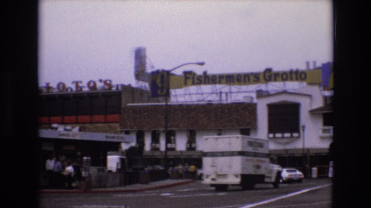 1973 San Francisco frame with Fishermen's Grotto signage above storefronts as a delivery truck and a white car travel through the waterfront street scene.