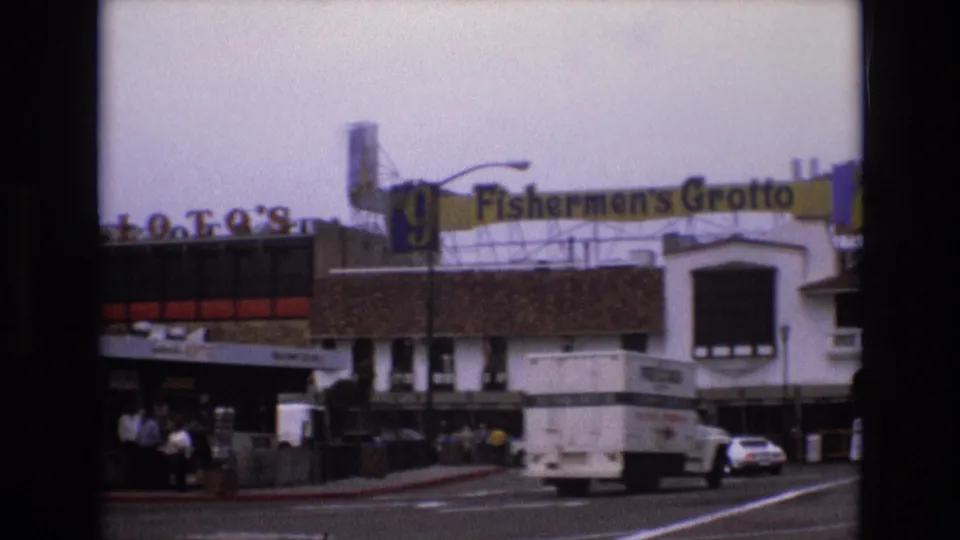 1973 San Francisco frame with Fishermen's Grotto signage above storefronts as a delivery truck and a white car travel through the waterfront street scene.