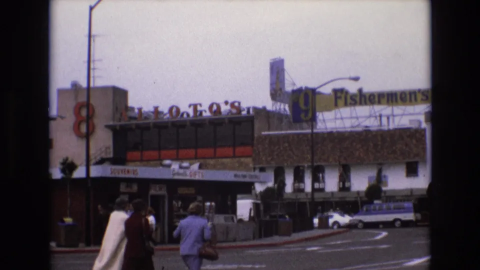 1973 San Francisco frame showing pedestrians in period clothing crossing the foreground near Fishermen's Grotto and neighboring tourist storefronts.
