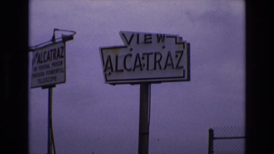 1973 San Francisco home-movie frame showing a roadside VIEW ALCATRAZ sign and a smaller sign advertising a telescope view of the former federal prison.