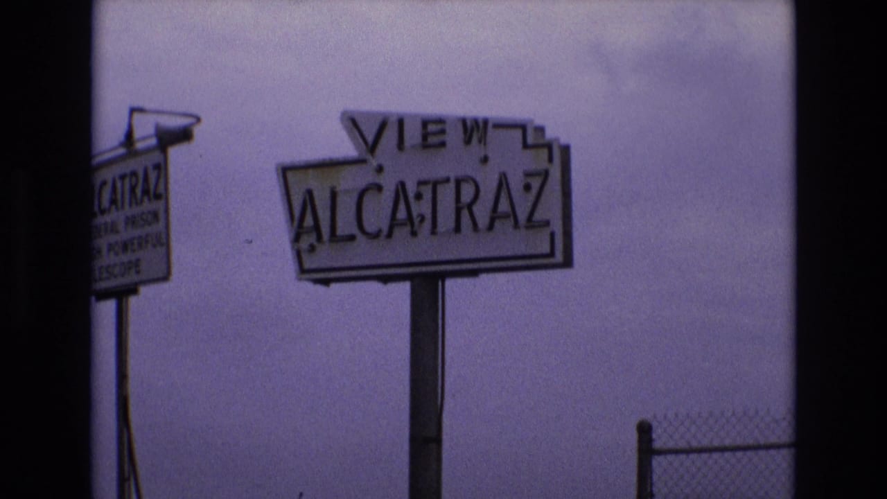 1973 San Francisco frame with the VIEW ALCATRAZ roadside sign centered, a partial telescope advertisement sign on the left, and chain-link fencing at the lower right.
