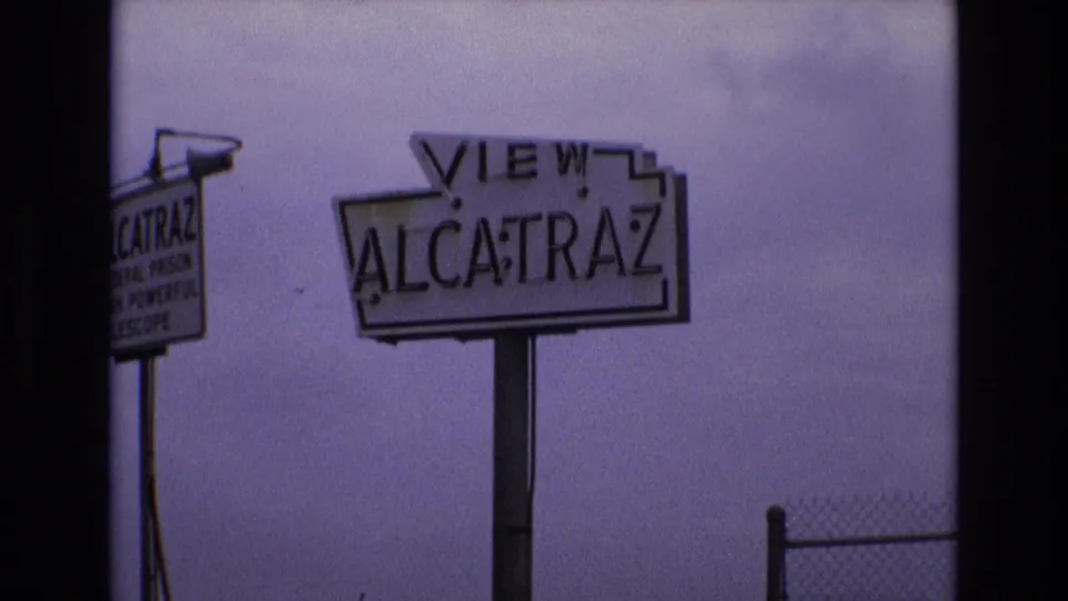 1973 San Francisco frame with the VIEW ALCATRAZ roadside sign centered, a partial telescope advertisement sign on the left, and chain-link fencing at the lower right.