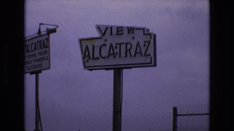1973 San Francisco home-movie frame holding on the VIEW ALCATRAZ sign, with a smaller Alcatraz telescope sign partly visible and a chain-link fence in the foreground.