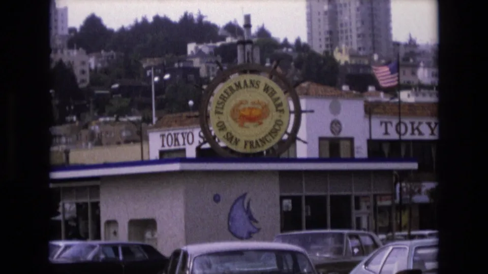 1973 San Francisco frame showing a Fisherman's Wharf commercial building with a circular crab sign, TOKYO storefront lettering, parked cars, an American flag, and hillside buildings in the background.