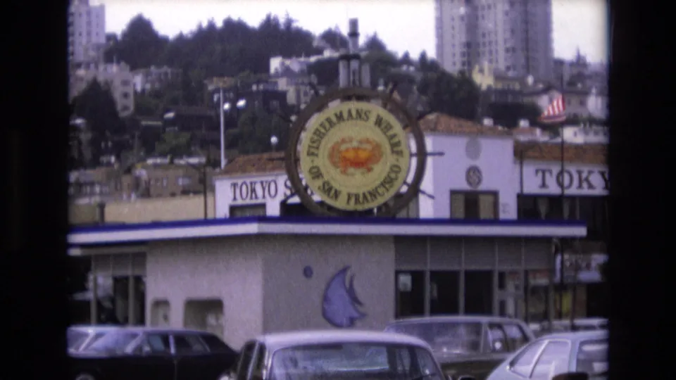 1973 San Francisco home-movie frame showing the circular Fisherman's Wharf crab sign, Tokyo restaurant lettering, parked cars, and hillside buildings behind the waterfront storefronts.
