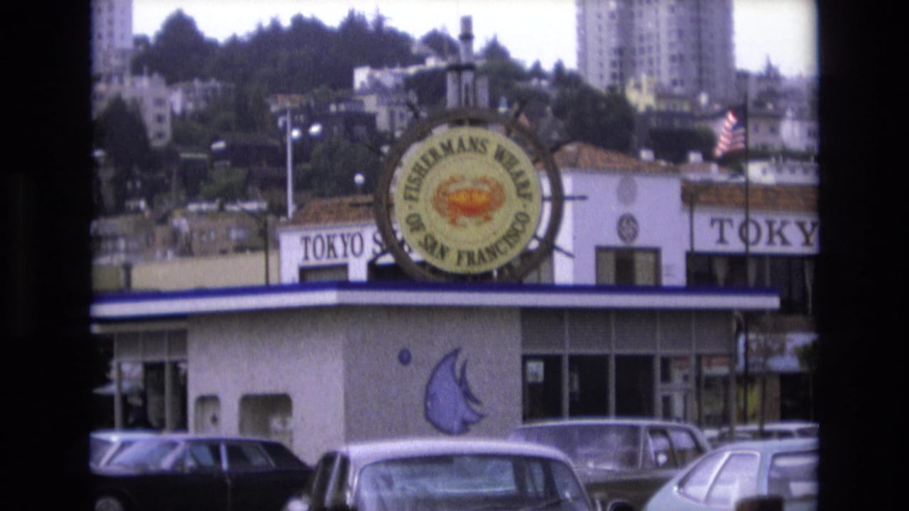 1973 San Francisco frame with Fisherman's Wharf signage centered above a low restaurant building, parked period cars below, and hillside apartments and trees in the background.