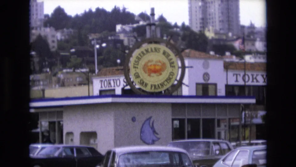 1973 San Francisco 8mm frame showing the Fisherman's Wharf crab sign, partial Tokyo restaurant sign, blue-trimmed storefront, parked automobiles, and hillside buildings beyond.