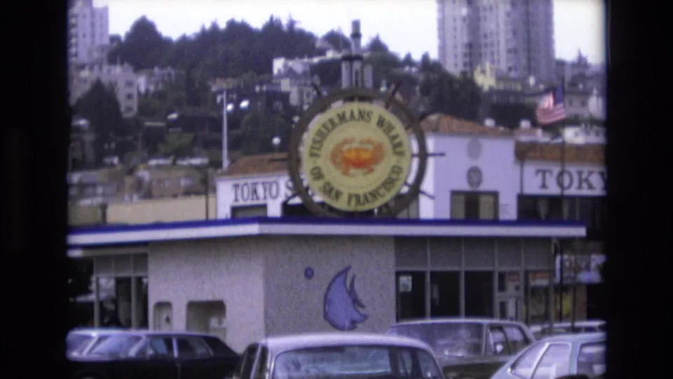 Final 1973 San Francisco frame holding on the Fisherman's Wharf sign and nearby restaurant facade, with parked cars in front and a wooded hillside with taller buildings behind.