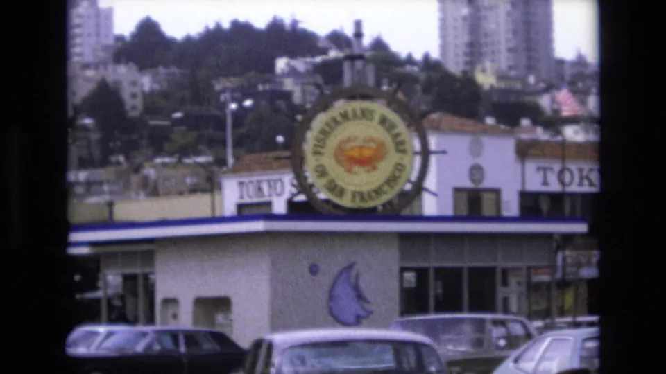 1973 San Francisco home-movie frame showing a Fisherman's Wharf facade with a circular crab-and-wheel sign, parked 1970s cars, and hillside buildings behind it.