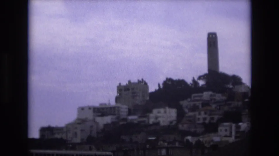 1973 San Francisco home-movie frame with Coit Tower on the hillside, clustered buildings below, and a waterfront railing along the lower edge.