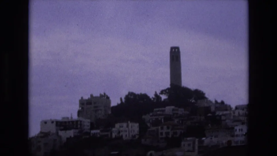 1973 San Francisco frame showing Coit Tower and hillside buildings in darker exposure, with trees and a cloudy sky visible across the skyline.