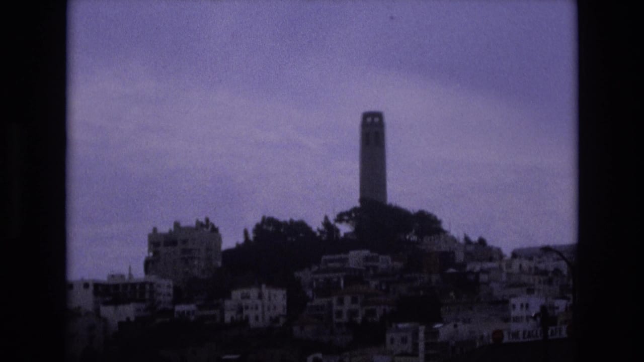 Coit Tower rises above a dark hillside and clustered residential buildings under a muted sky.