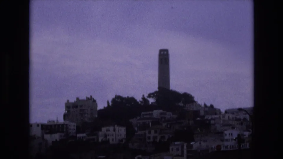 1973 San Francisco 8mm frame showing Coit Tower above Telegraph Hill-style residential buildings, with dark trees, soft focus, and visible film-frame edges.
