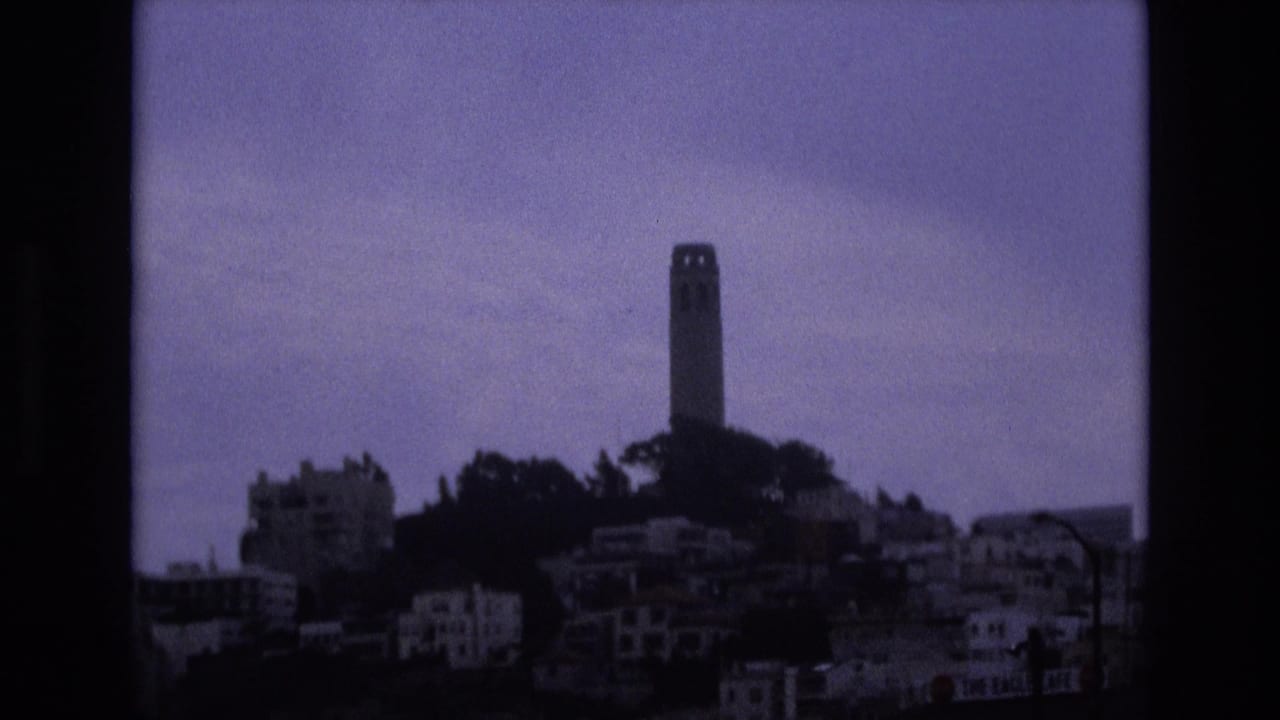 Distant 1973 home-movie view of Coit Tower overlooking San Francisco hillside housing, framed by dark 8mm borders and soft purple-blue color.