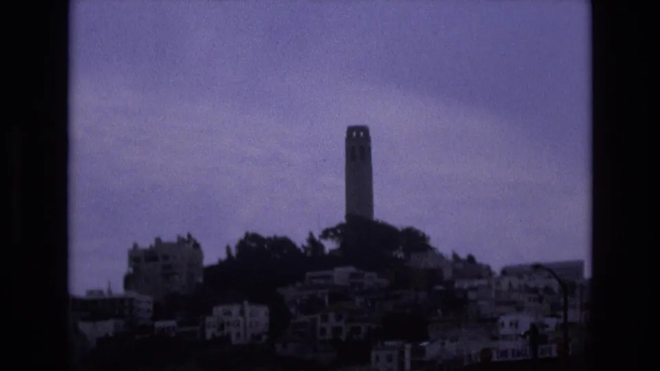 Distant 1973 home-movie view of Coit Tower overlooking San Francisco hillside housing, framed by dark 8mm borders and soft purple-blue color.