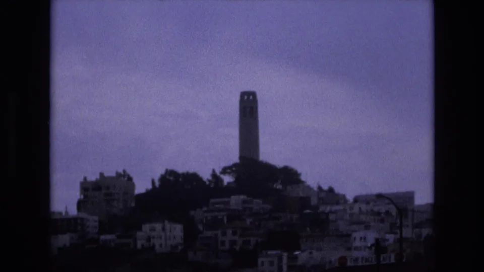 Soft 8mm frame from San Francisco in 1973 showing Coit Tower centered above a dark hill, clustered buildings, and a muted sky.
