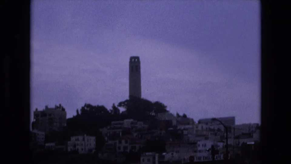 1973 San Francisco home-movie still showing Coit Tower above dense hillside residences, with dark border areas, grain, and cool color shift.