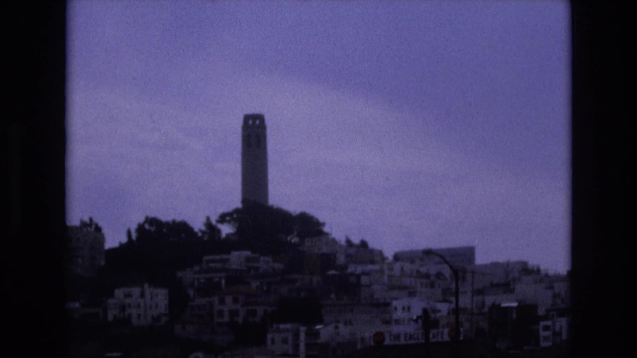 Coit Tower above a dark hillside of San Francisco buildings under an overcast sky.