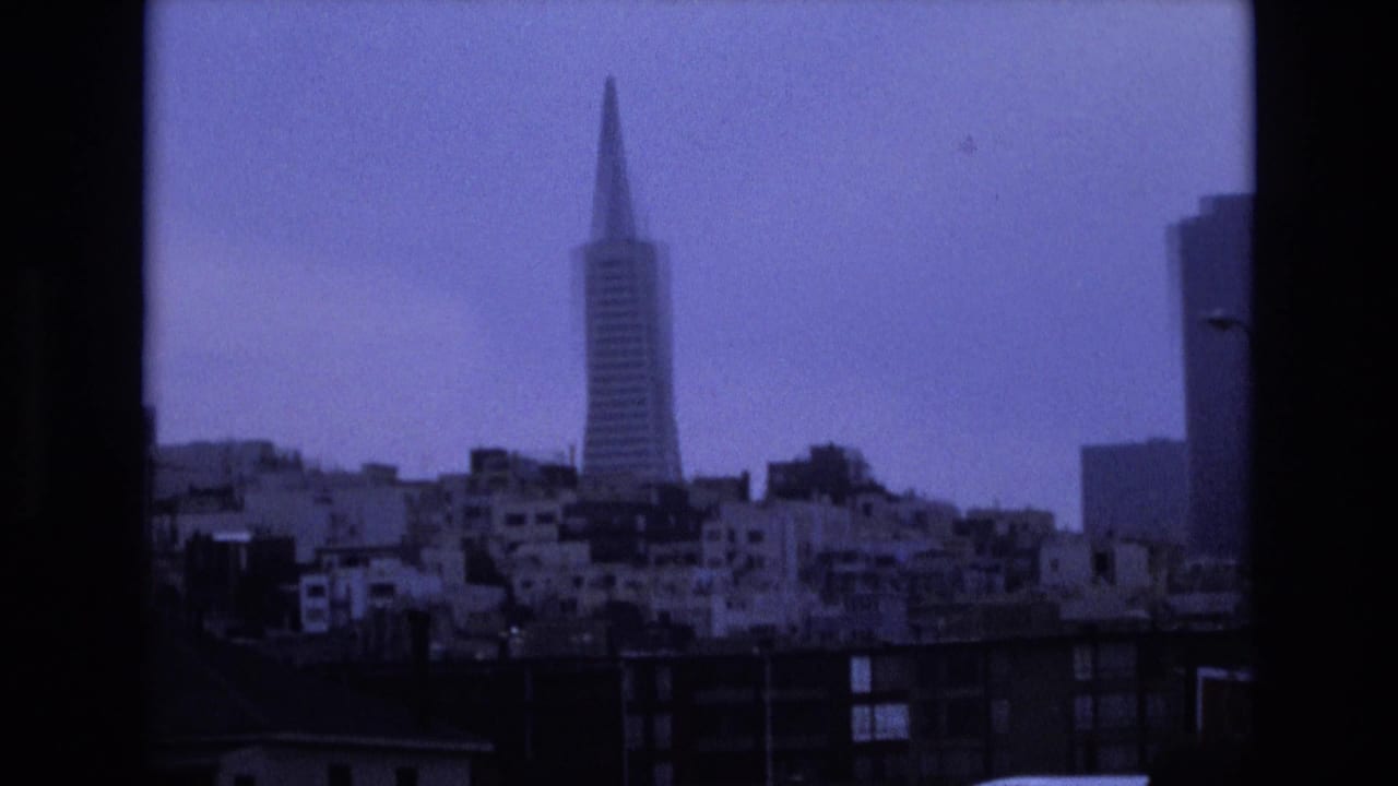 1973 San Francisco skyline view with the Transamerica Pyramid centered above dense residential buildings, seen through dark window-like frame edges on blue-tinted 8mm film.