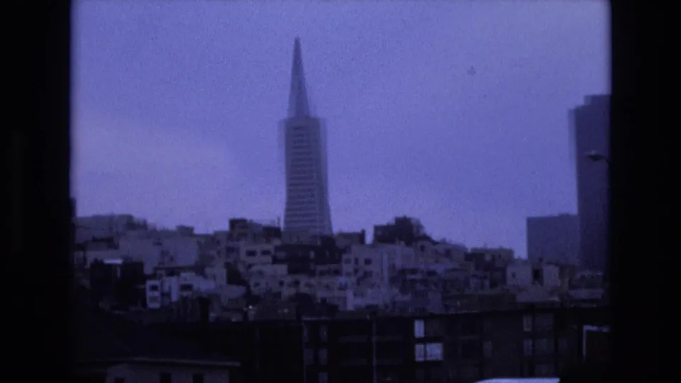 1973 San Francisco skyline view with the Transamerica Pyramid centered above dense residential buildings, seen through dark window-like frame edges on blue-tinted 8mm film.
