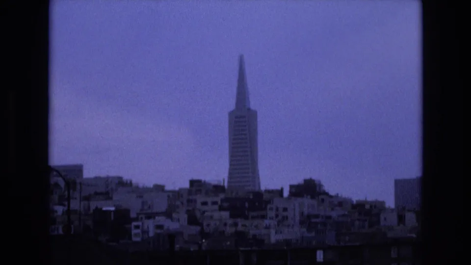 1973 San Francisco home-movie frame with the Transamerica Pyramid prominent over layered rooftops and apartment buildings beneath a pale blue-gray sky.