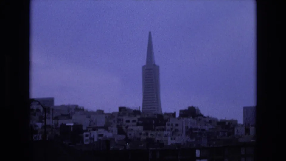 1973 San Francisco skyline still with the Transamerica Pyramid set behind clustered buildings, a dark roofline in the foreground, and a cool blue-purple 8mm color cast.