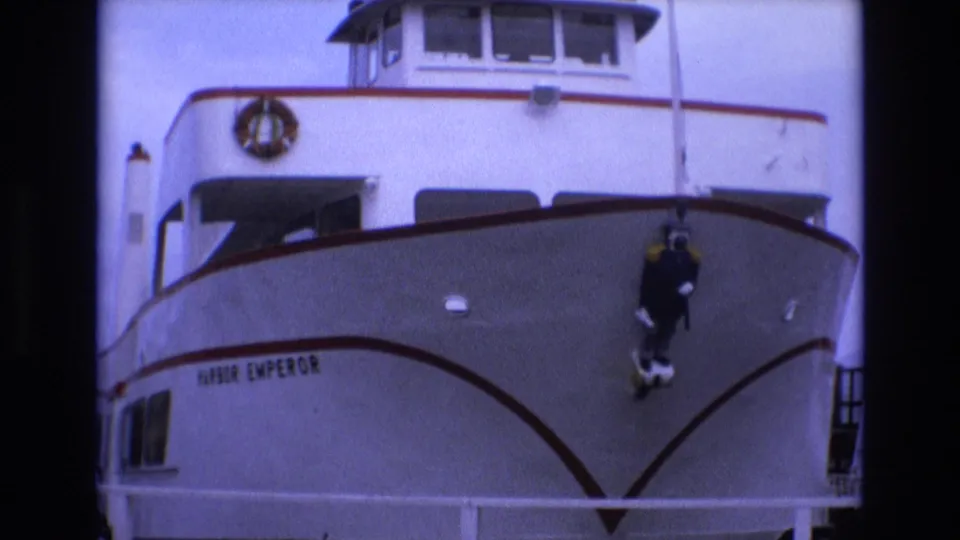1973 San Francisco 8mm frame showing the white Harbor Emperor bow with red trim, wheelhouse windows, a life ring, and a dark diver figure suspended against the hull.